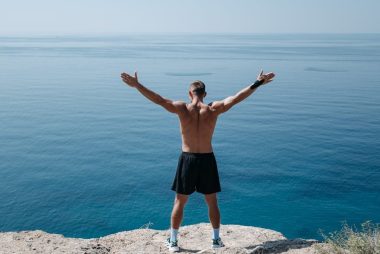 A shirtless man stands on a cliff with open arms, embracing freedom and the vast ocean.